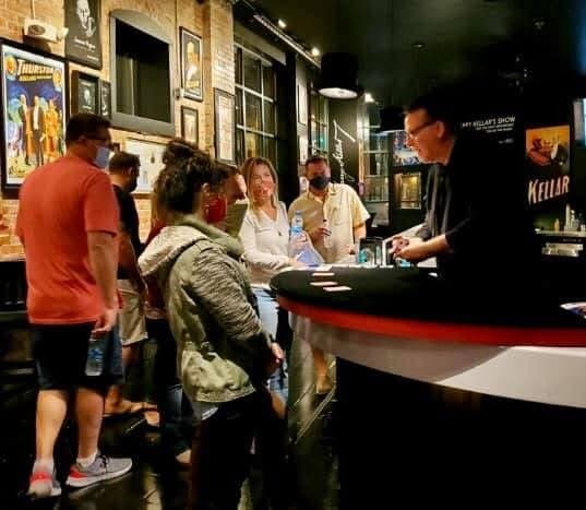 Eric Thompson delights a small gathering with an intimate magic performance, drawing curious onlookers around his table as he works his comedic charm in what appears to be a cozy venue setting.