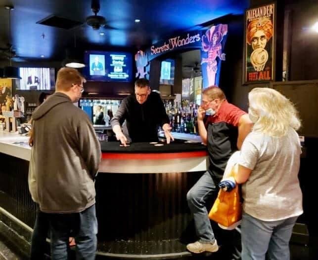 Eric Thompson mesmerizes a small crowd at what appears to be a bar or restaurant venue, working his magic up close with an intimate audience gathered around.