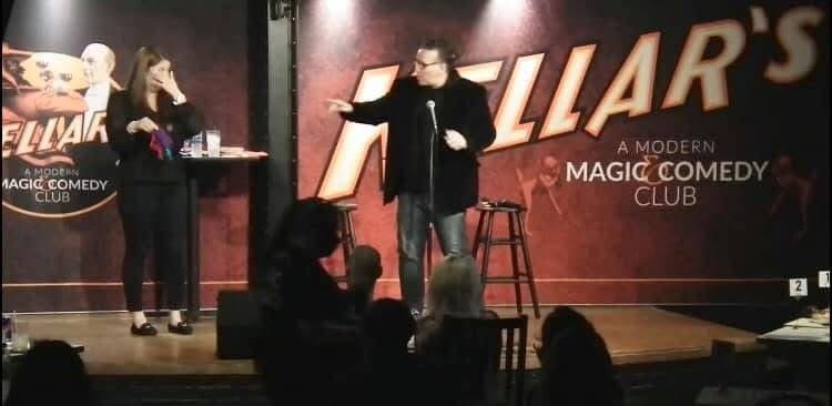 Eric Thompson commands the stage at a modern comedy club, delivering his signature blend of magic and laughs to an intimate audience gathered around the performance area.