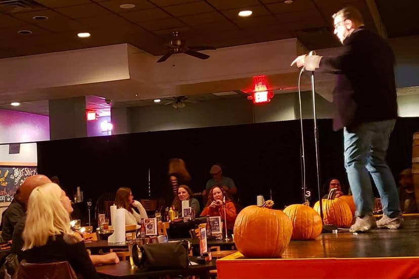 Eric Thompson towers above the crowd at what appears to be a festive autumn-themed restaurant event, working his magic while surrounded by decorative pumpkins and an engaged audience enjoying dinner and drinks.