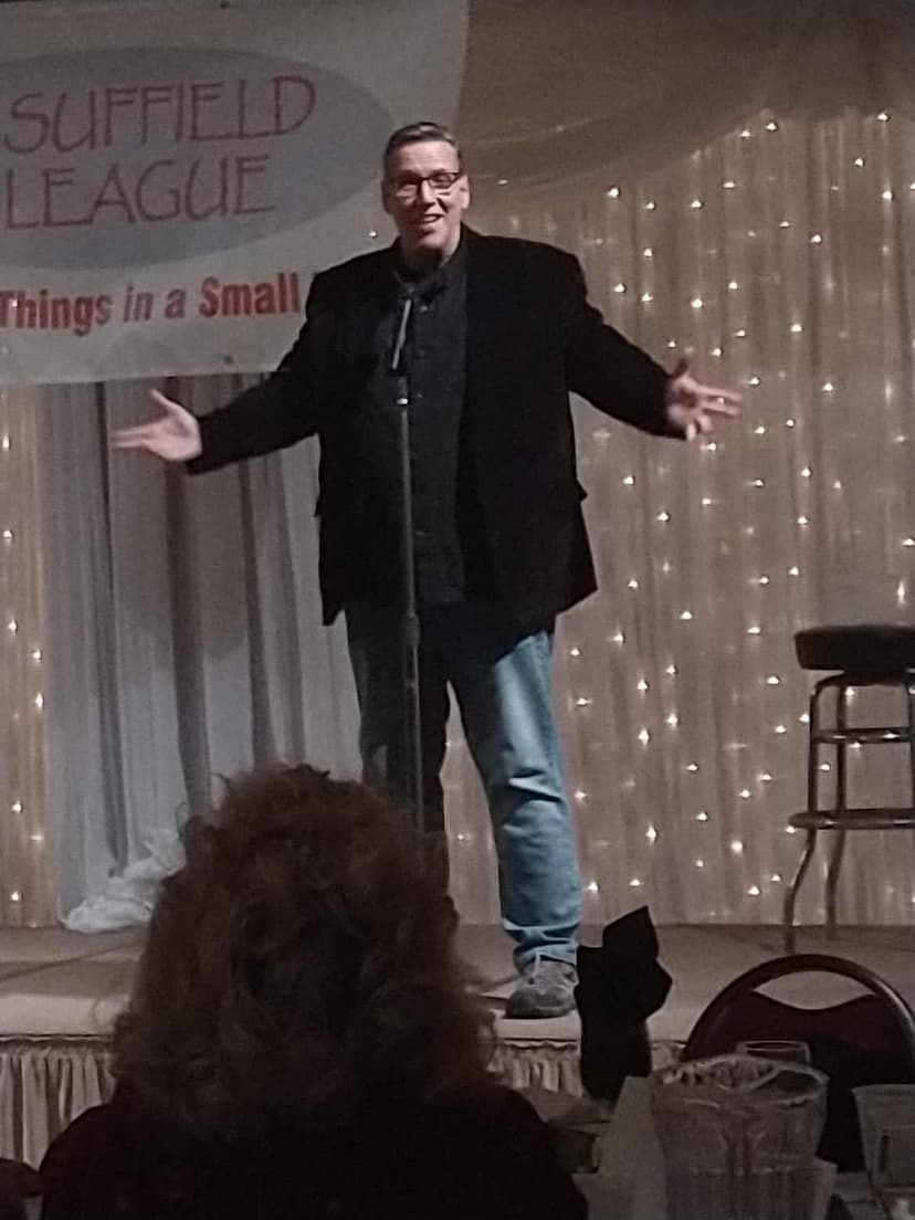 Eric Thompson commands the stage with his signature animated style, arms spread wide as he delivers his blend of comedy and magic to an appreciative audience at what appears to be a cozy venue.