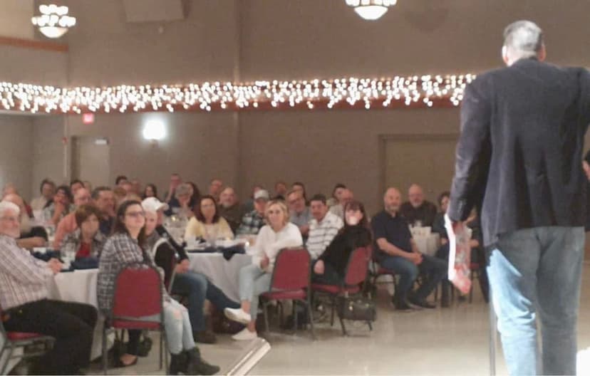 Eric Thompson commands the room at what appears to be a festive banquet hall event, with a captivated audience of dozens hanging on his every word as twinkling lights create the perfect ambiance for an unforgettable evening of entertainment.