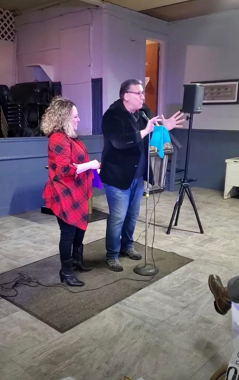 Eric Thompson captivates his audience during what appears to be an intimate performance, gesturing expressively while a woman in a festive red plaid coat watches with delight from nearby.