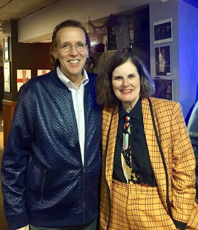 Eric Thompson shares a warm moment with a colleague backstage, both dressed to impress in stylish blazers that show off their distinctive fashion sense.