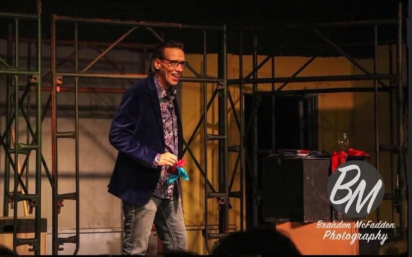 Eric Thompson takes center stage in his signature blazer and patterned shirt, working his magic on what appears to be a theater audience with his trademark blend of comedy and illusion.