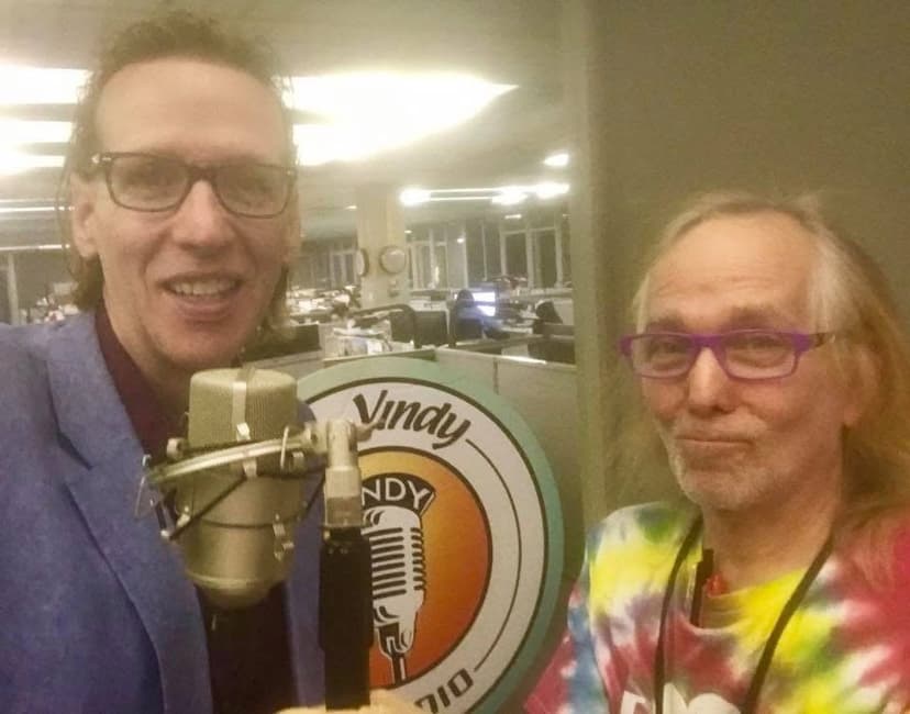 Eric Thompson takes a break from his comedy routine to pose with a fellow performer in what appears to be a radio studio, complete with professional microphone and station branding visible in the background.