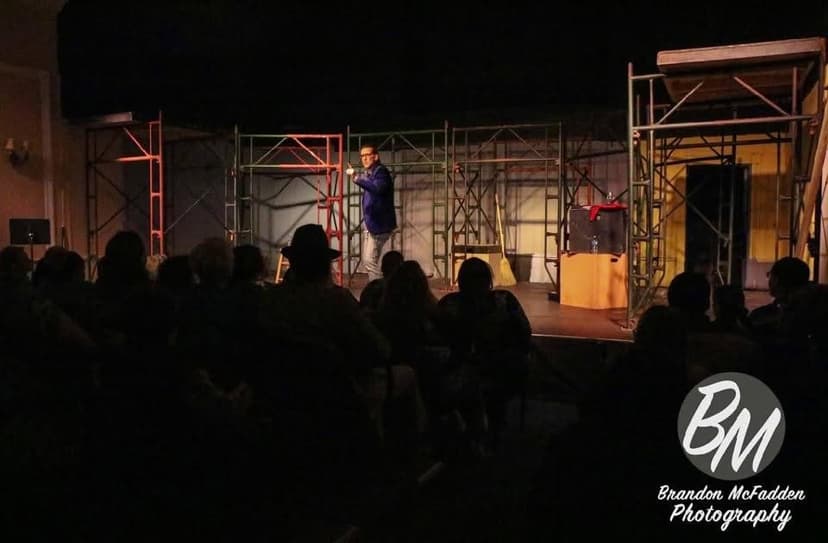 Eric Thompson commands the stage at what appears to be a theater performance, working his magic with an engaged audience silhouetted in the foreground. The industrial-style set design creates a perfect backdrop for Eric's blend of comedy and illusion.
