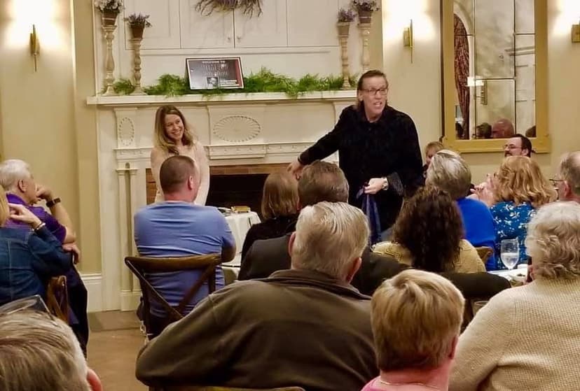 Eric Thompson captivates an intimate audience gathered around a cozy fireplace, delivering his signature blend of comedy and charm in what appears to be a private home or upscale venue.