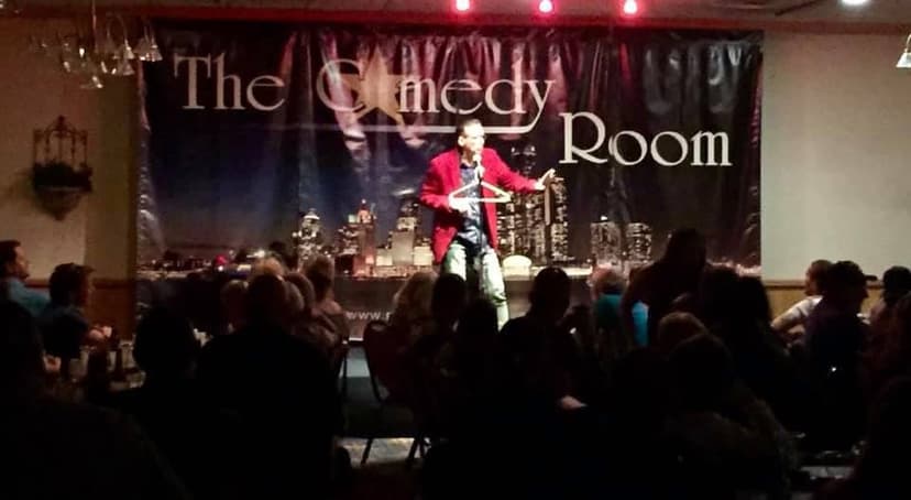 Eric Thompson commands the stage in his signature red jacket, delivering laughs to a packed house at The Comedy Room with the city skyline backdrop setting the perfect scene for an unforgettable night of entertainment.