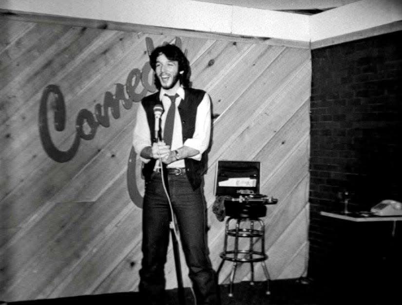 Eric Thompson takes the stage at what appears to be a comedy club, microphone in hand and ready to deliver his signature blend of humor and magic to an eager audience.