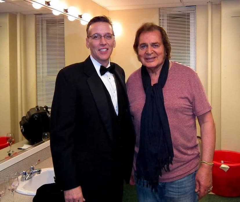 Eric Thompson shares a backstage moment with a fellow entertainer, both all smiles after what looks like a successful show - Eric's formal tuxedo contrasting perfectly with his friend's casual post-performance look.