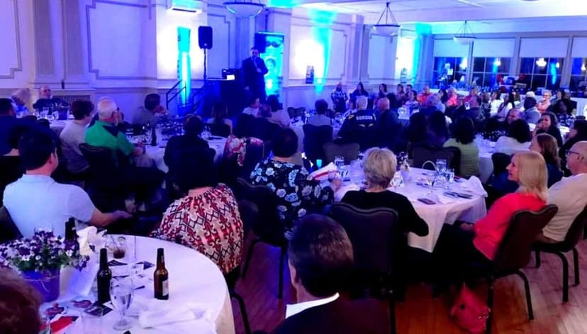 Eric Thompson commands attention from a packed banquet hall filled with elegantly dressed guests, the blue stage lighting creating a perfect atmosphere for an evening of comedy and magic.