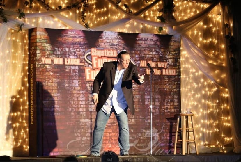 Eric Thompson brings his signature energy to the stage at what appears to be a comedy club, striking a dynamic pose under warm string lights with a classic brick wall backdrop setting the perfect atmosphere for laughs.