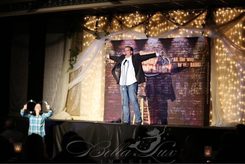Eric Thompson commands the stage with arms outstretched, delivering his signature blend of comedy and magic under twinkling lights while an enthusiastic audience member cheers him on from below.
