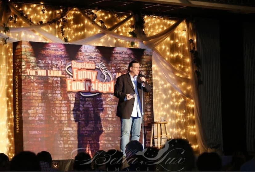 Eric Thompson takes the stage under a canopy of twinkling lights, bringing his signature warmth and humor to what looks like an intimate comedy club setting with that classic brick wall backdrop.