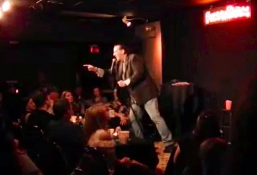 Eric Thompson commands the stage at an intimate comedy club, working his magic with a captivated audience hanging on his every word in the moody stage lighting.