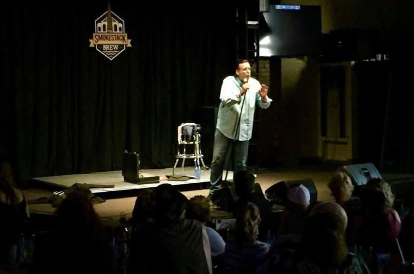 Eric Thompson commands the stage at what appears to be a comedy club or brewery venue, microphone in hand as he delivers his signature blend of humor to an engaged audience seated below.