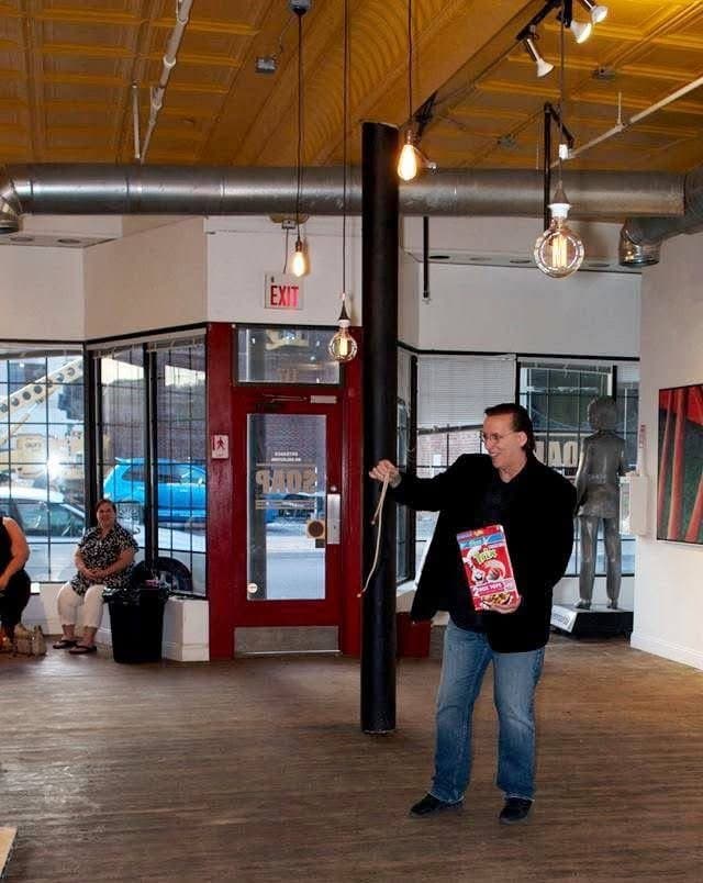 Eric Thompson works the room with his signature charm, holding what appears to be a colorful prop while entertaining guests at this loft-style venue with its industrial chic atmosphere.