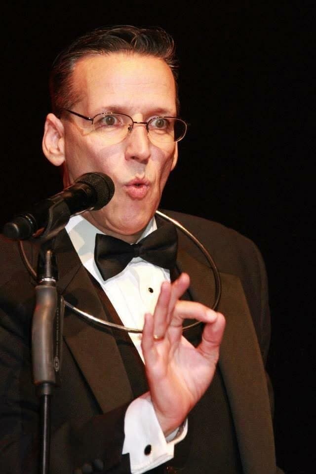 Eric Thompson commands the stage in classic formal wear, his animated gestures and expressive face showing why he's been captivating audiences since the early 1980s. The seasoned entertainer appears mid-performance, likely weaving together his signature blend of comedy and magic for a delighted crowd.